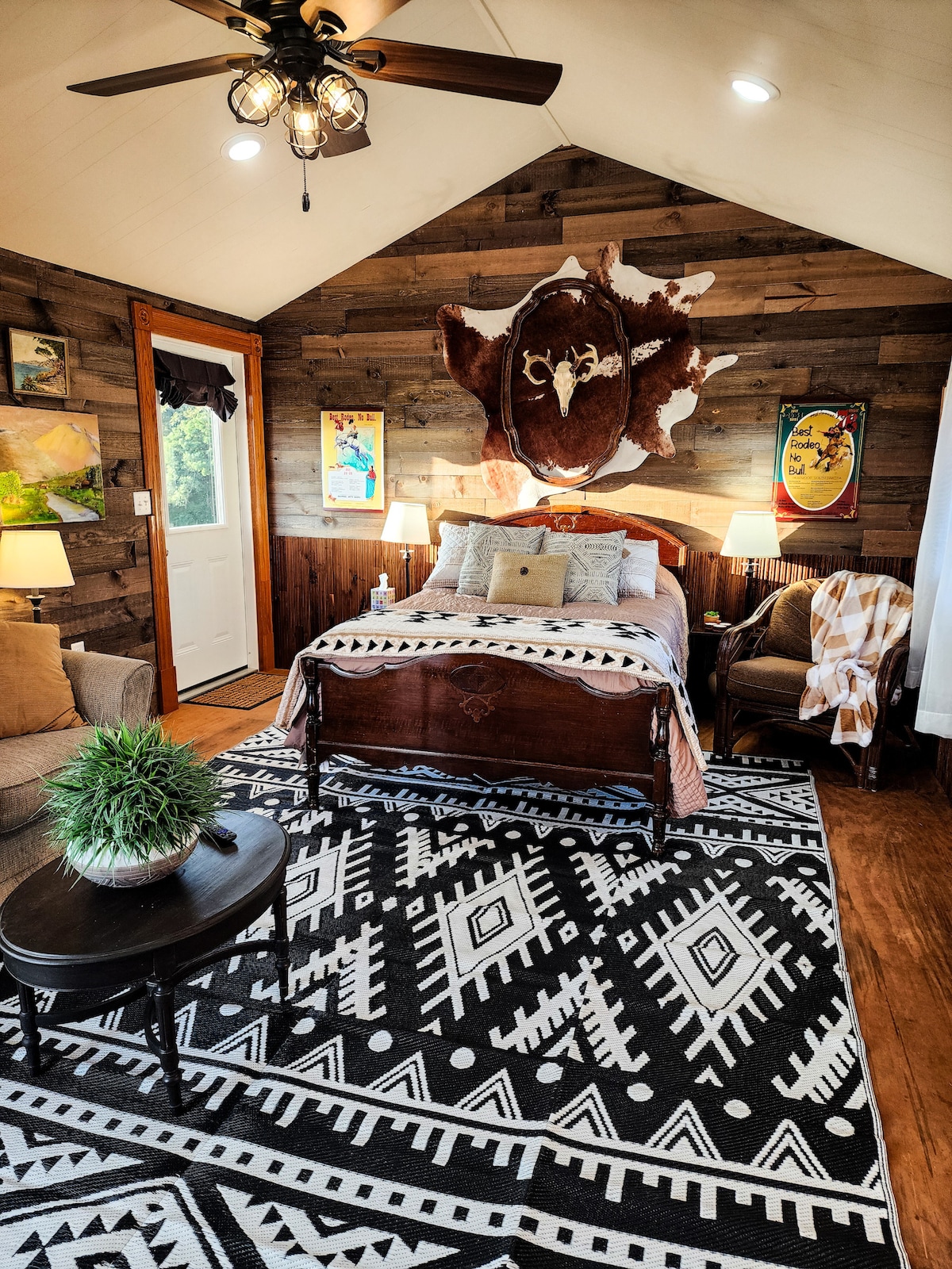 A cozy sleeping area is featured, showcasing a full-sized bed with decorative pillows and a soft blanket. The rustic wooden walls are complemented by artwork and a mounted hide. A comfortable chair and a small table enhance the inviting ambiance, illuminated by soft light fixtures.