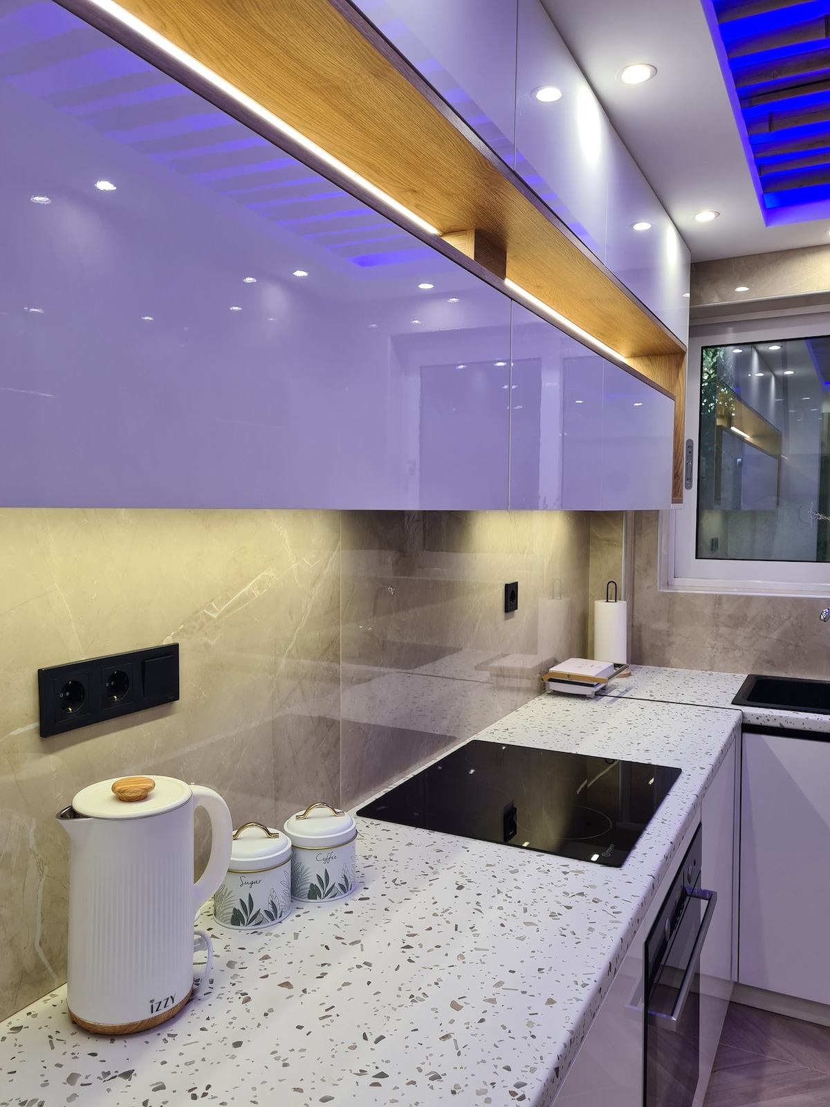 A modern kitchen is displayed featuring smooth, glossy cabinets and a spacious countertop with a speckled design. Under-cabinet lighting enhances the sleek surfaces. An electric cooktop is positioned on the counter, with a kettle and decorative containers nearby. A large window provides natural light.