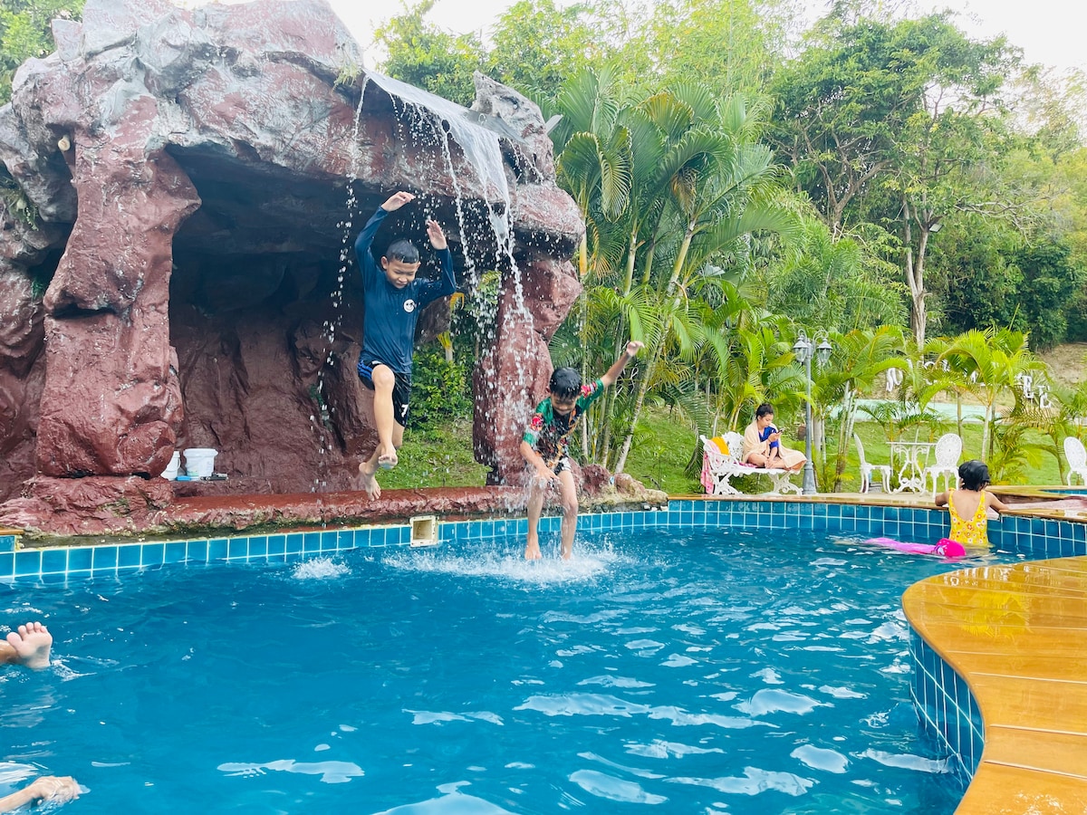 A swimming pool is surrounded by lush greenery, featuring a waterfall cascading from a rocky structure. Two individuals are mid-air, jumping into the pool, while others relax nearby. Several chairs are positioned along the pool's edge, inviting leisure and enjoyment.