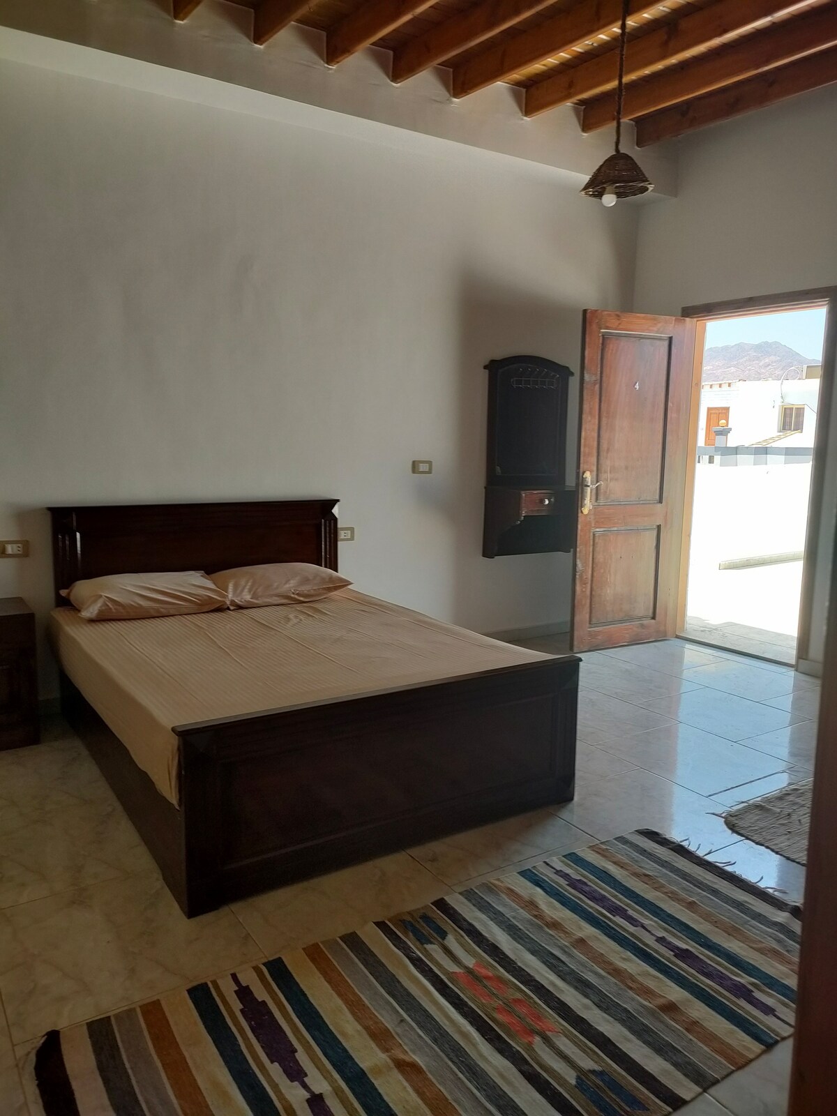 A room features a wooden bed with a neutral-toned bedspread and accent pillows. Natural light enters through an open door leading to an outdoor area, while a rug with varying colors adds warmth to the tiled floor.