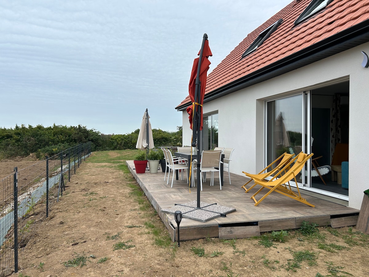 An outdoor terrace features seating for several guests, including a shaded table and chairs. A bright yellow deck chair is positioned beside the entrance, with an umbrella offering protection from the sun. The surrounding area is flat with grass and greenery visible in the distance.