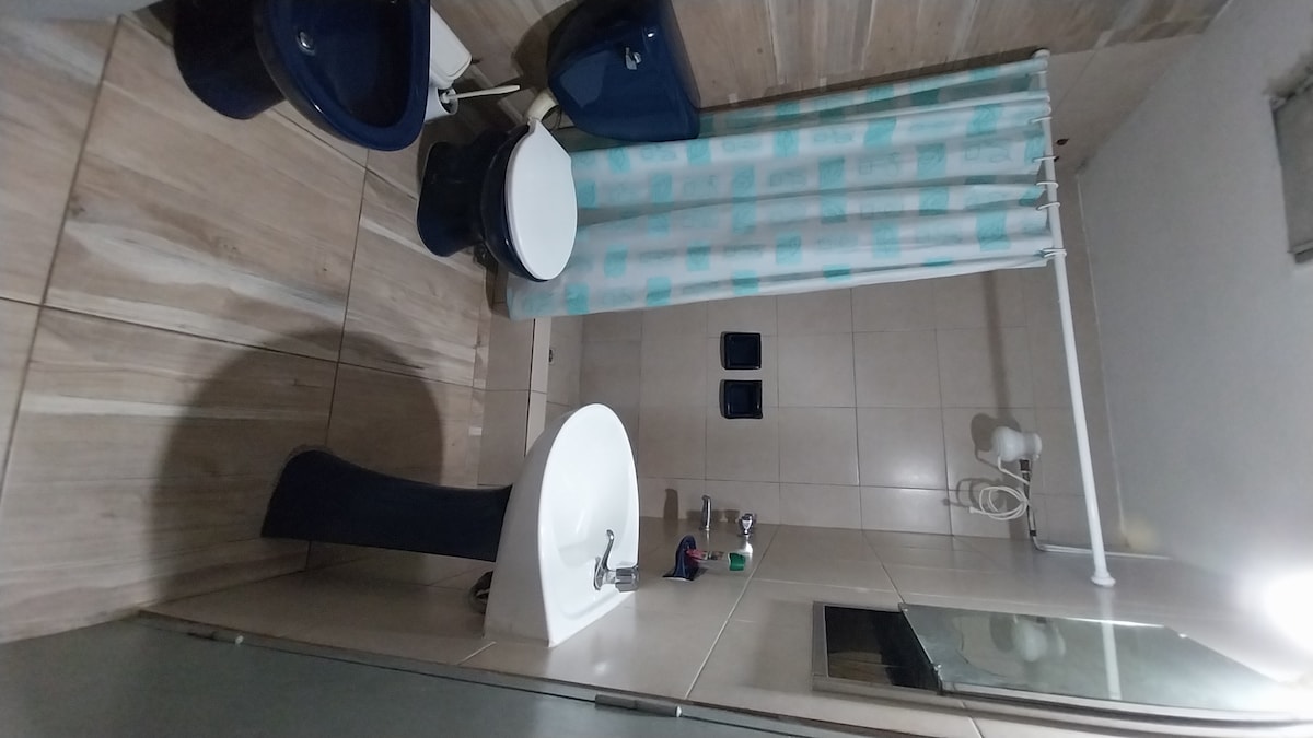 The bathroom layout features modern fixtures, including a toilet and a sink with a sleek design. A shower area is visible, separated by a glass wall. A laundry shower curtain in light blue and white adds a subtle touch of color. Neutral tiles adorn the floor and walls.