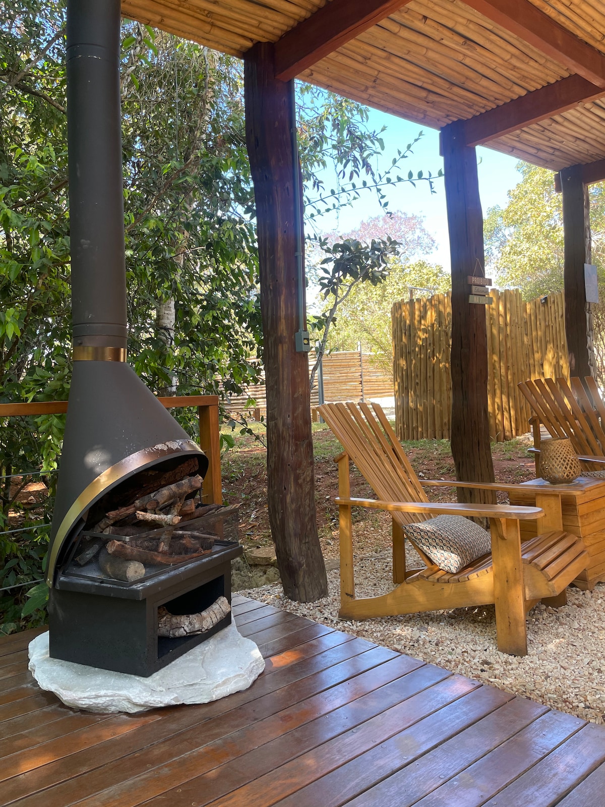 A cozy outdoor seating area features two wooden lounge chairs positioned beside a stone fire pit. The space is shaded by a wooden pergola, surrounded by lush greenery, creating a peaceful environment for relaxation.