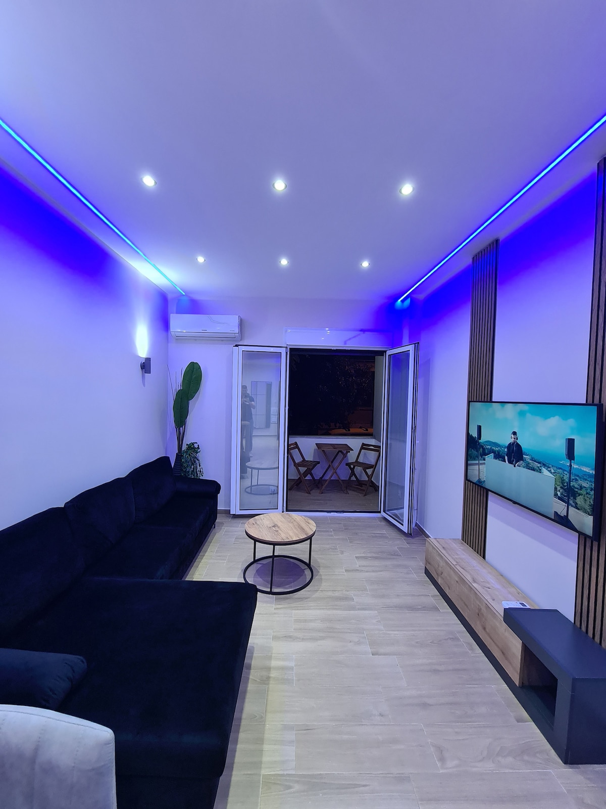The living area is furnished with a dark sectional sofa facing a wall-mounted television. Soft blue LED lighting enhances the ambiance. A small, circular coffee table occupies the center, and double doors open to a balcony, allowing easy access to outdoor seating.
