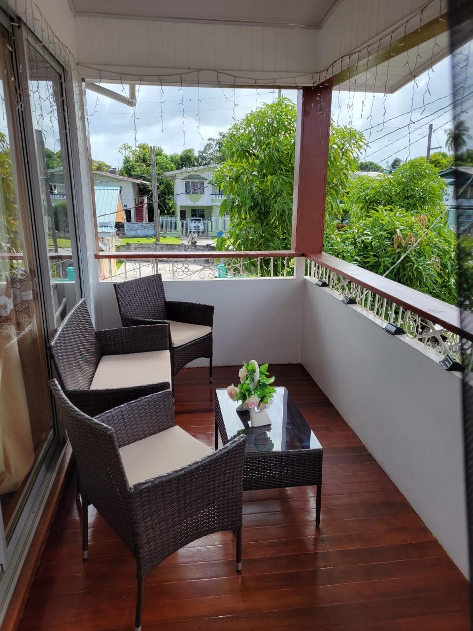 A covered balcony is furnished with four wicker chairs and a glass coffee table. Potted plants and greenery are present, contributing to a refreshing atmosphere. Natural light filters through the nearby glass doors, illuminating the wooden flooring.