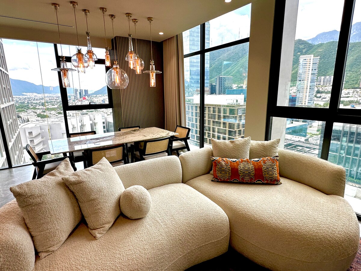A modern living area is highlighted by a curvy, plush sofa adorned with decorative cushions. Large floor-to-ceiling windows offer expansive views of the surrounding city and mountains. A stylish dining table is positioned nearby, illuminated by contemporary light fixtures hanging above.