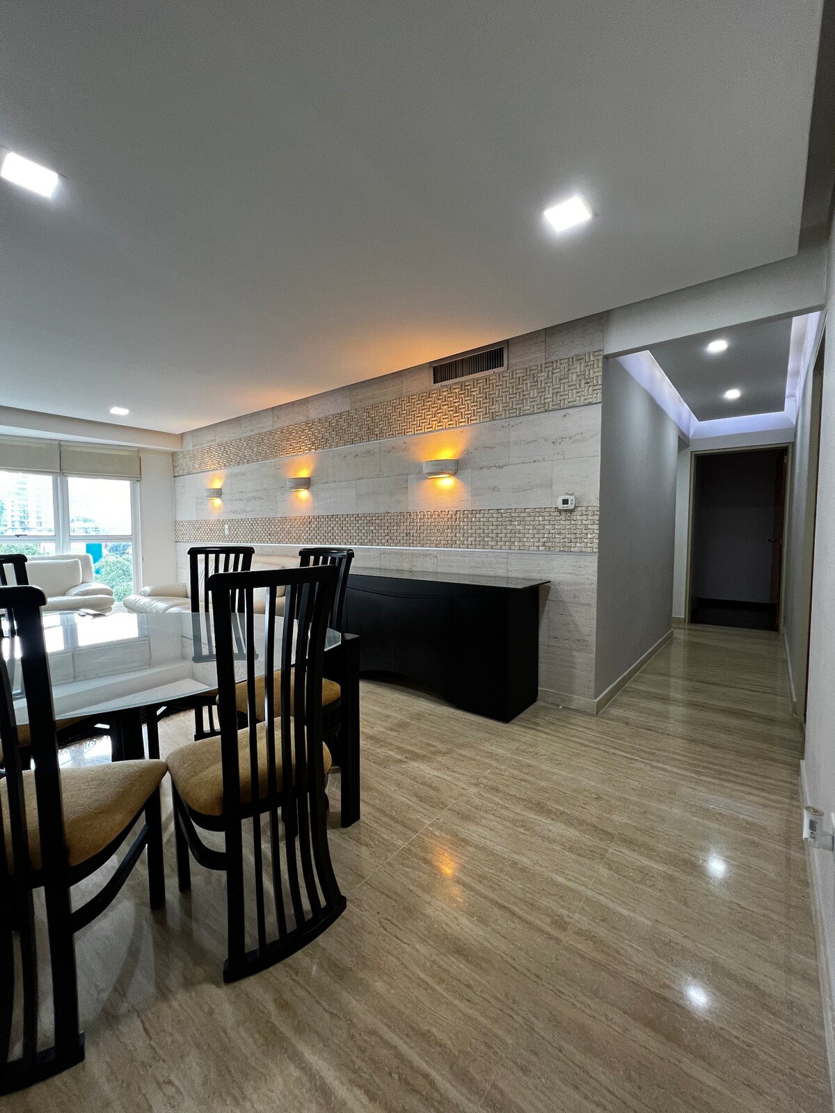 A well-lit dining area features a glass-top table surrounded by six black-styled chairs. The walls display a subtle stone texture, complemented by warm sconces that provide ambient lighting. A hallway is visible in the background, leading to additional spaces in the apartment.