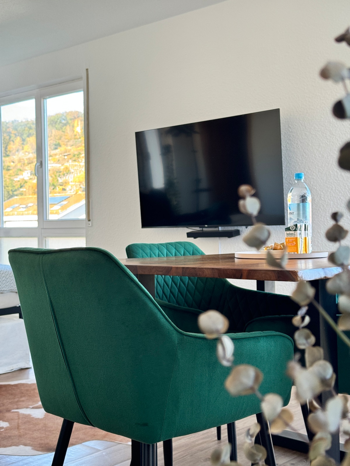 A dining area is highlighted with a wooden table and two plush green chairs. A flat-screen TV mounted on the wall is visible, alongside a water bottle and a small drink placed on the table. Natural light filters through a nearby window.