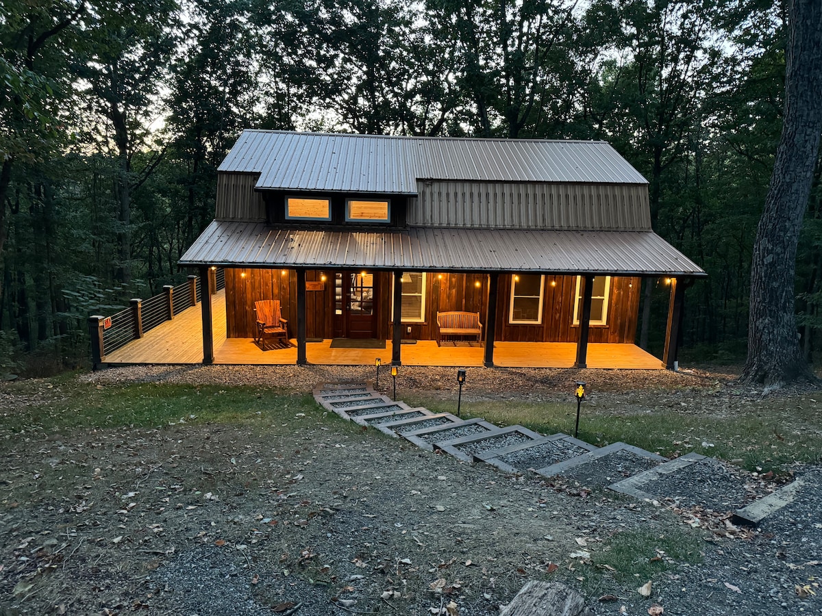 A rustic barn-style cabin, featuring a copper metal roof and large front porch illuminated by warm lighting. Surrounded by trees, a gravel walkway leads to the entrance, inviting guests to explore the natural beauty of the 20-acre property.