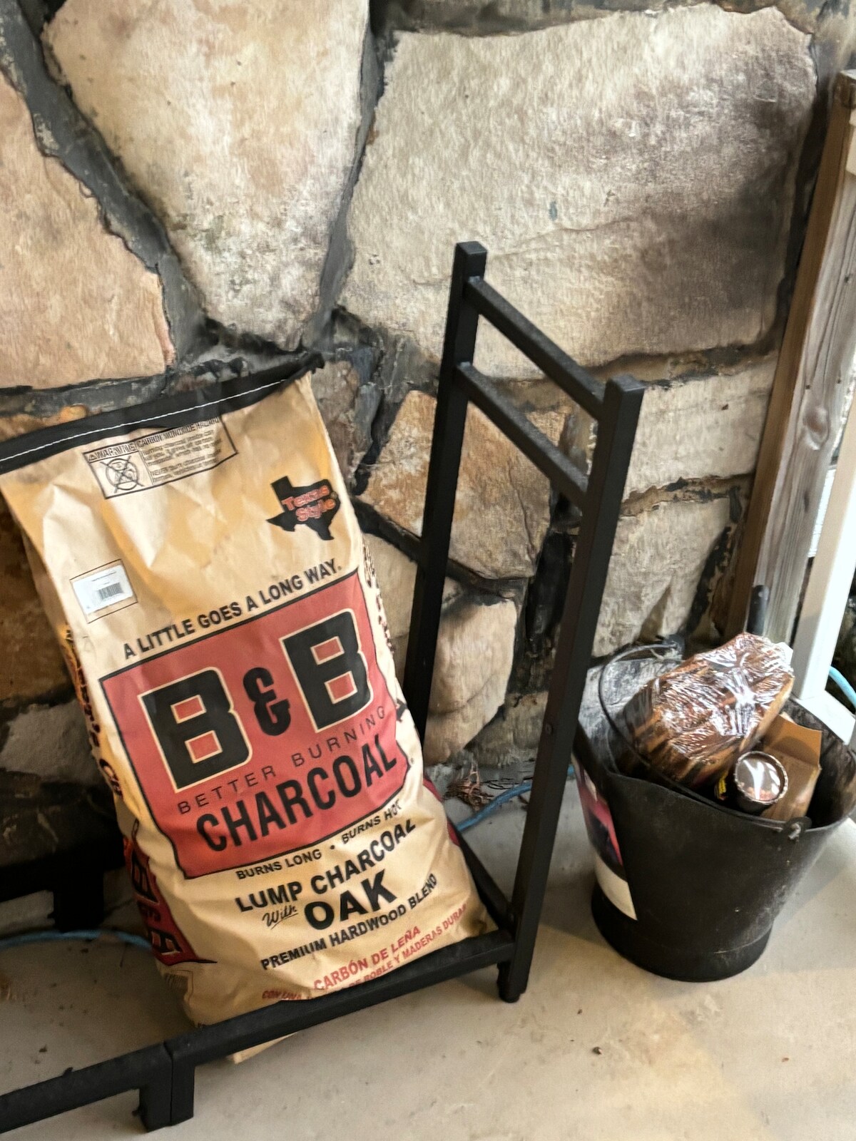 A charcoal bag labeled B&B sits upright next to a black metal holder. A small bucket nearby contains firewood, all positioned against a textured stone wall, which adds rustic charm to the setting.