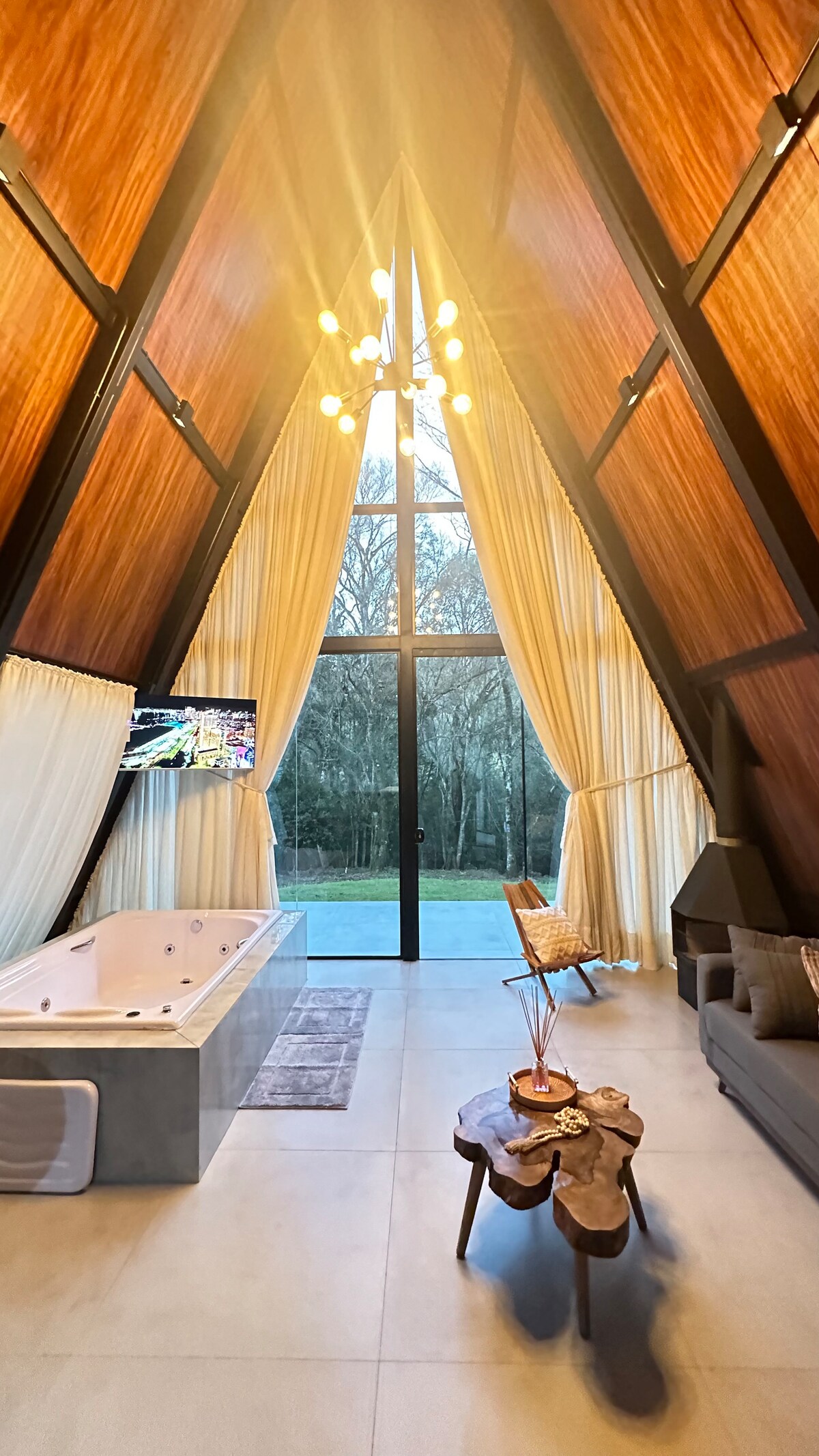 The interior features a high, vaulted ceiling with wooden beams and large windows allowing natural light to fill the space. A stylish jacuzzi is set beside a cozy seating area, which includes a sofa and a decorative wooden table. Soft curtains frame the windows, enhancing the serene ambiance.