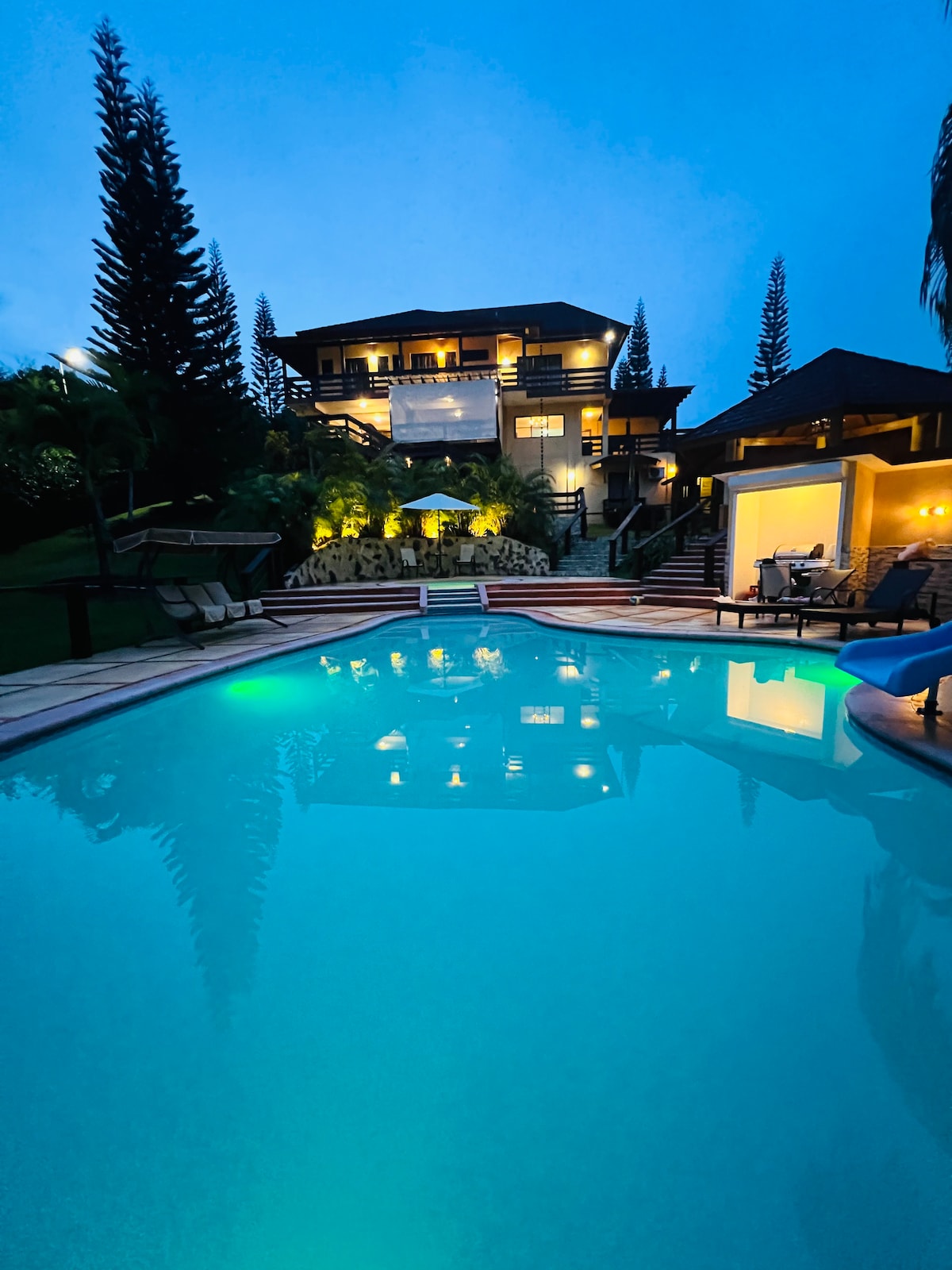 A serene evening scene features a well-lit villa overlooking a clear swimming pool. The inviting water reflects the lights from the villa, surrounded by tall trees and lush greenery that enhances the tranquil atmosphere.