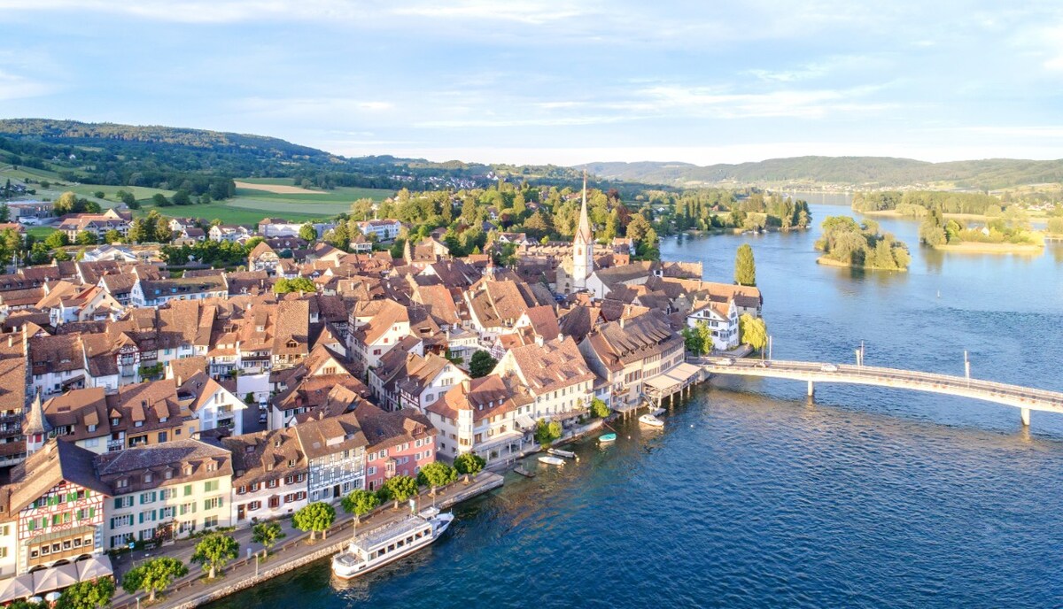 An aerial view captures the charming village of Stein am Rhein, characterized by a mix of rustic buildings with terracotta roofs. The banks of the Rhine River feature lush greenery and a bridge connecting each side, while boats are seen docked along the waterfront.