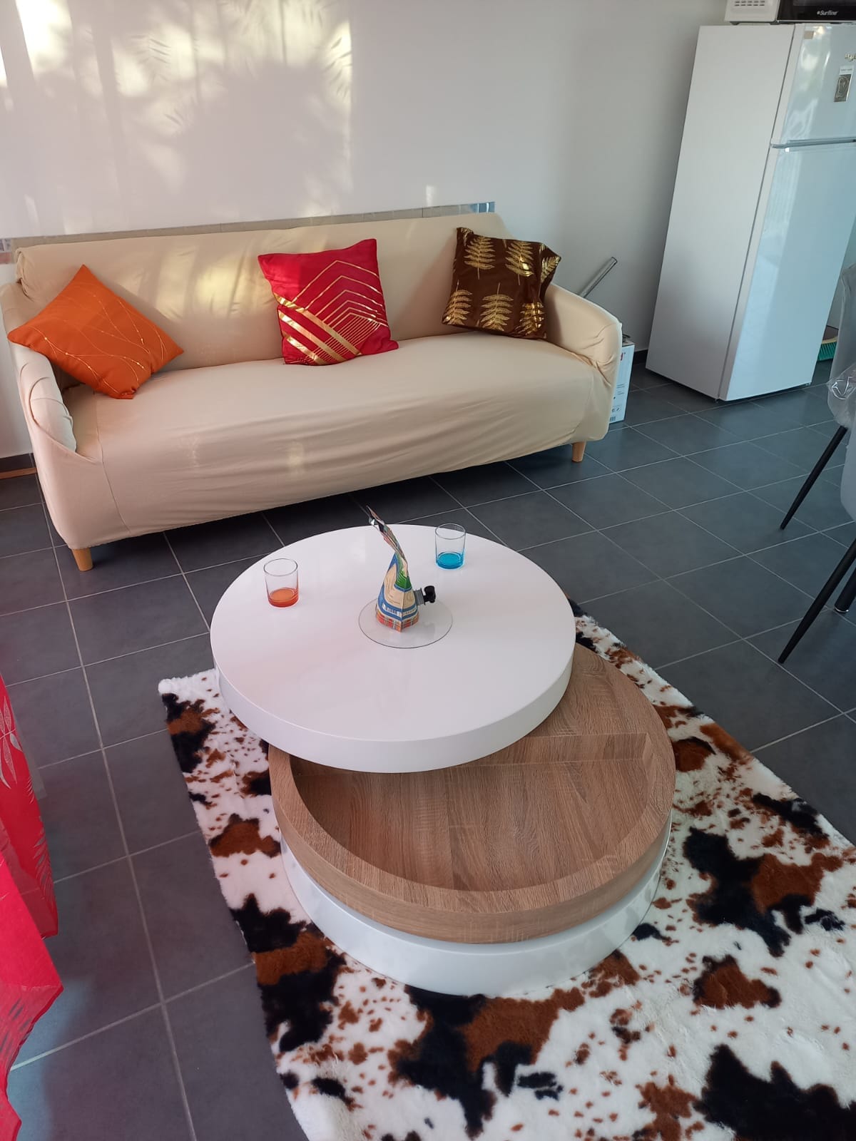 A living area is presented with a light-colored sofa adorned with decorative cushions. A round, dual-layer coffee table sits in the center on a patterned rug. A white refrigerator is visible in the background, contributing to the bright and spacious feel of the room.