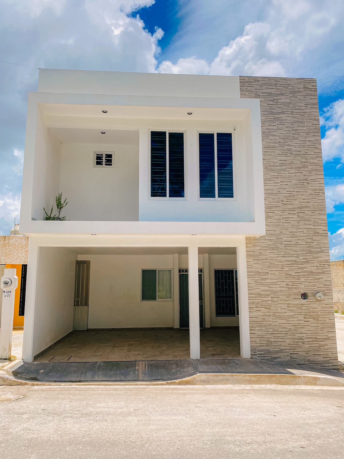 A modern two-story structure features a combination of smooth white walls and stone accents. Large windows allow ample natural light, while a covered parking area is present at the front. The clear sky and clouds create a bright backdrop for the building.