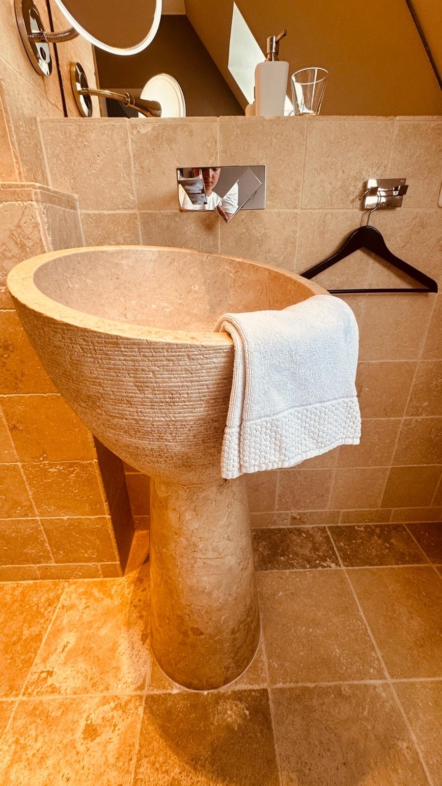 A natural stone sink with a textured surface is featured, supported by a cylindrical base. A neatly folded towel is hung over the rim. The surrounding walls are adorned with light-colored stone tiles, and a modern mirror reflects the warm lighting from above.