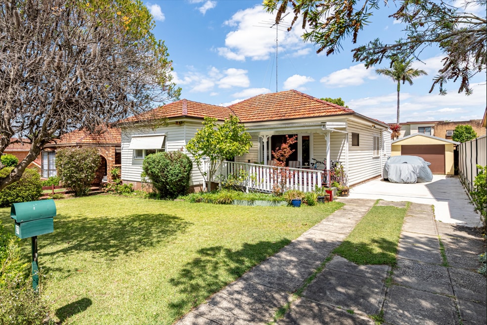 A single-story house is set in a neatly landscaped yard, featuring a covered front porch and a pathway leading to the entrance. Lush greenery and colorful plants enhance the outdoor space, while a large tree provides shade. A driveway extends to a garage at the rear.