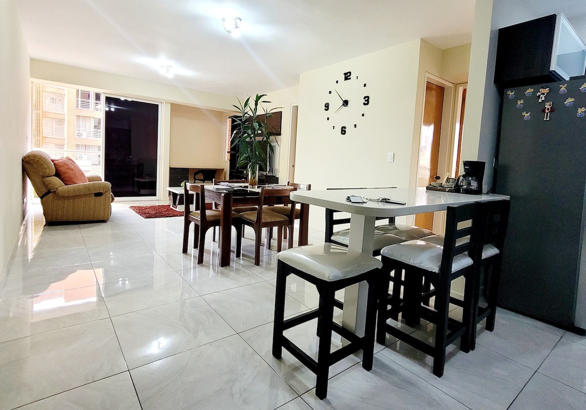 An open-plan living space is featured, showcasing a dining area with a rectangular table surrounded by chairs, along with a kitchen bar. A cozy seating area is present in the background, complemented by a large clock on the wall and light pouring in from expansive glass doors.