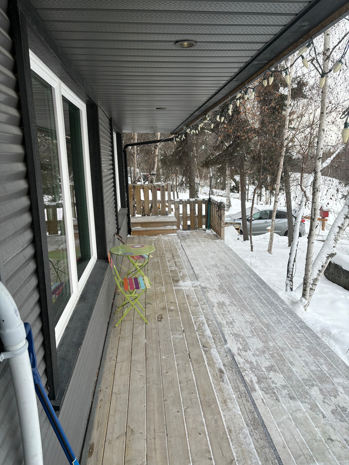 A private front deck is visible, featuring wooden flooring and outdoor seating in vivid green and striped colors. Snow covers the ground, and trees provide a natural backdrop. A wooden fence and glimpses of the surrounding area are also included.