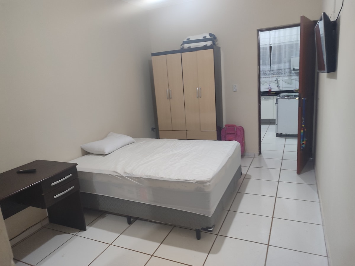 A simple bedroom is furnished with a spacious bed positioned against a neutral wall. A desk with drawers is situated nearby. A closet stands against the opposite wall, while a doorway leads to a kitchen area, visible in the background. Tile flooring contributes to the room's design.