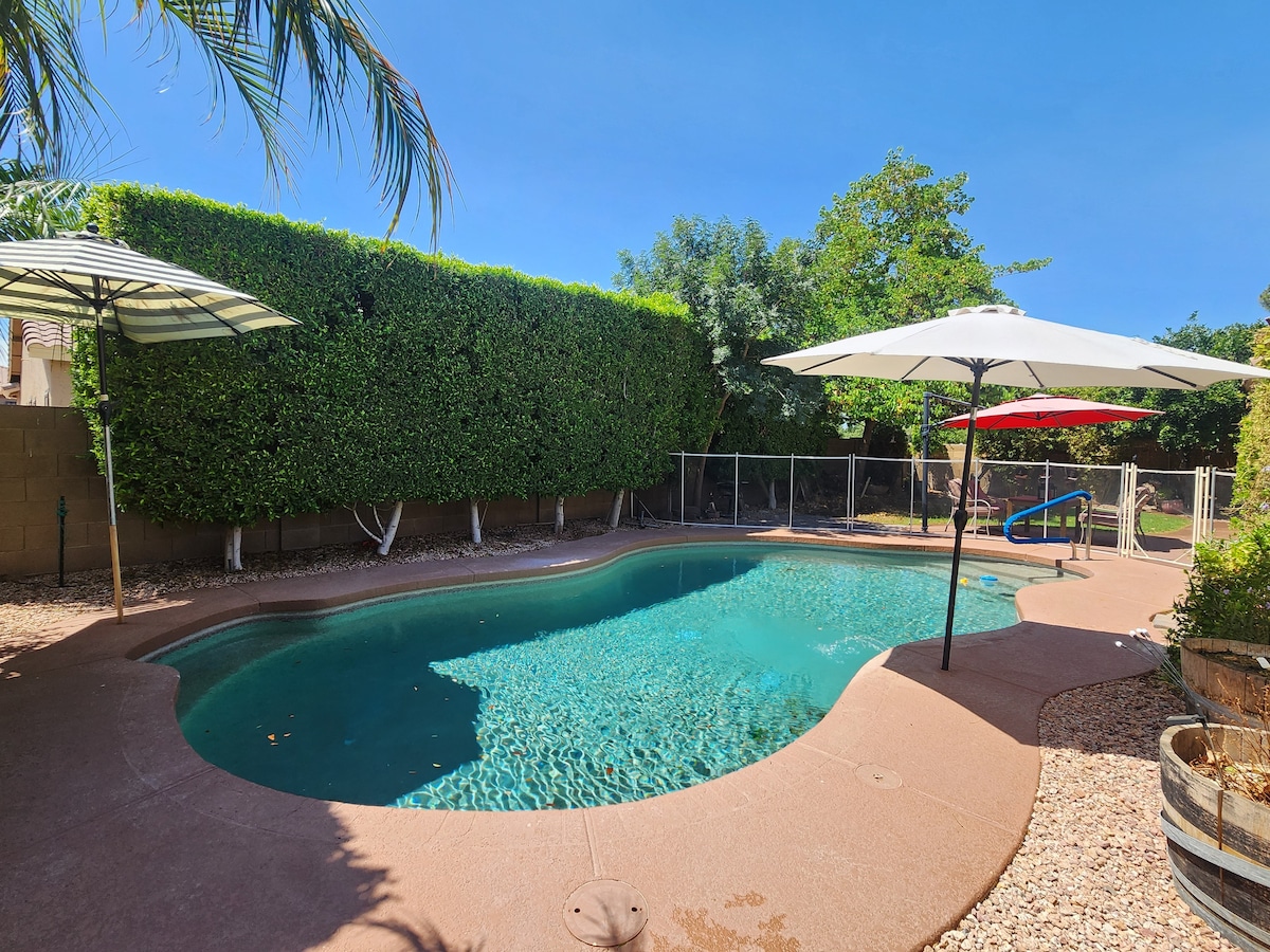 A private pool area is surrounded by lush green hedges and features two sun umbrellas. A clear blue pool reflects the sky above, with smooth stones lining the perimeter. Nearby, a fenced section provides safety, enhancing the outdoor experience.