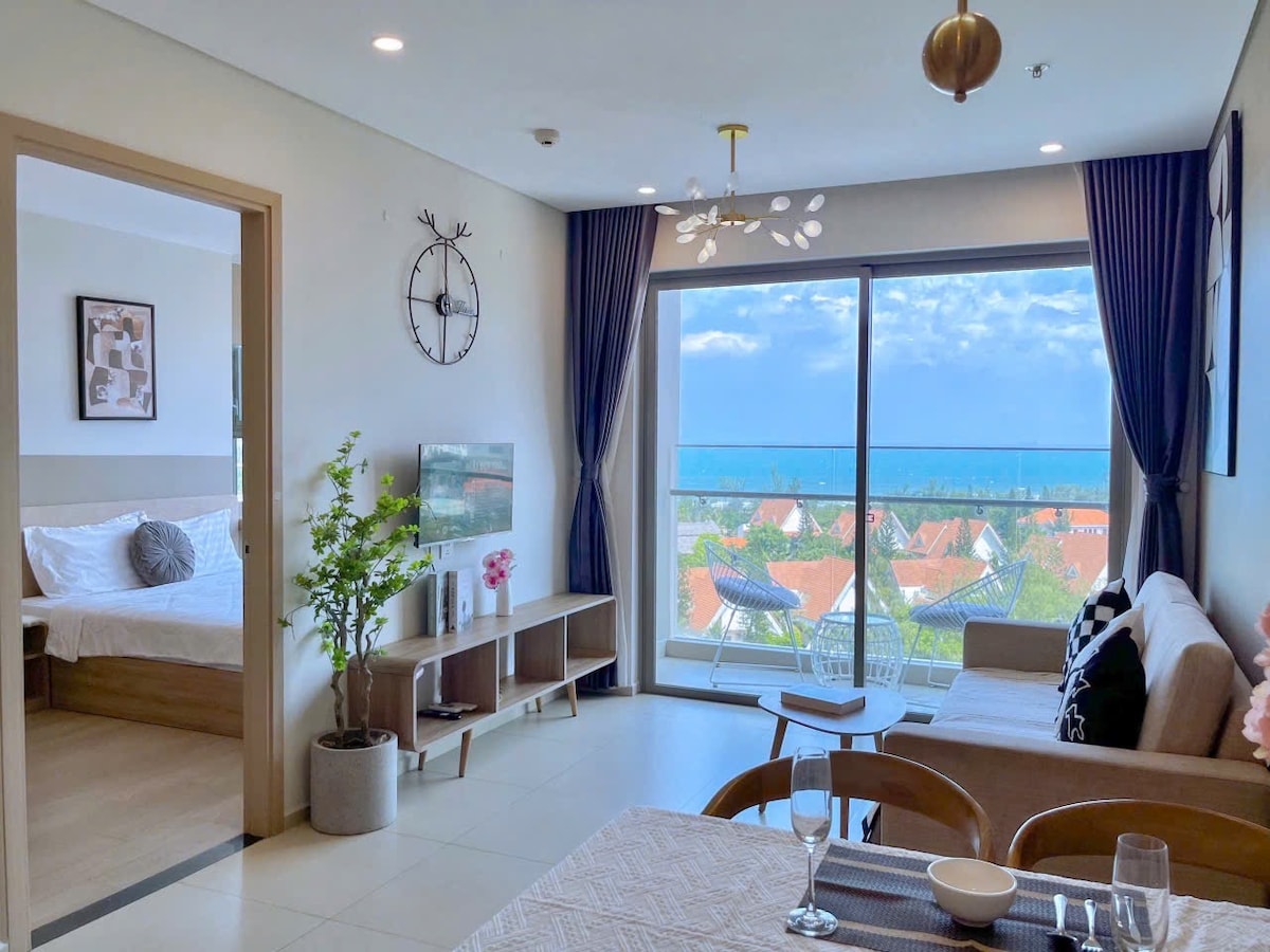 A bright living area features a large window with a view of the surrounding landscape. A modern sofa is positioned alongside a wooden coffee table. A dining area with simple table settings contributes to the inviting space. The adjacent room displays a glimpse of a spacious bed.