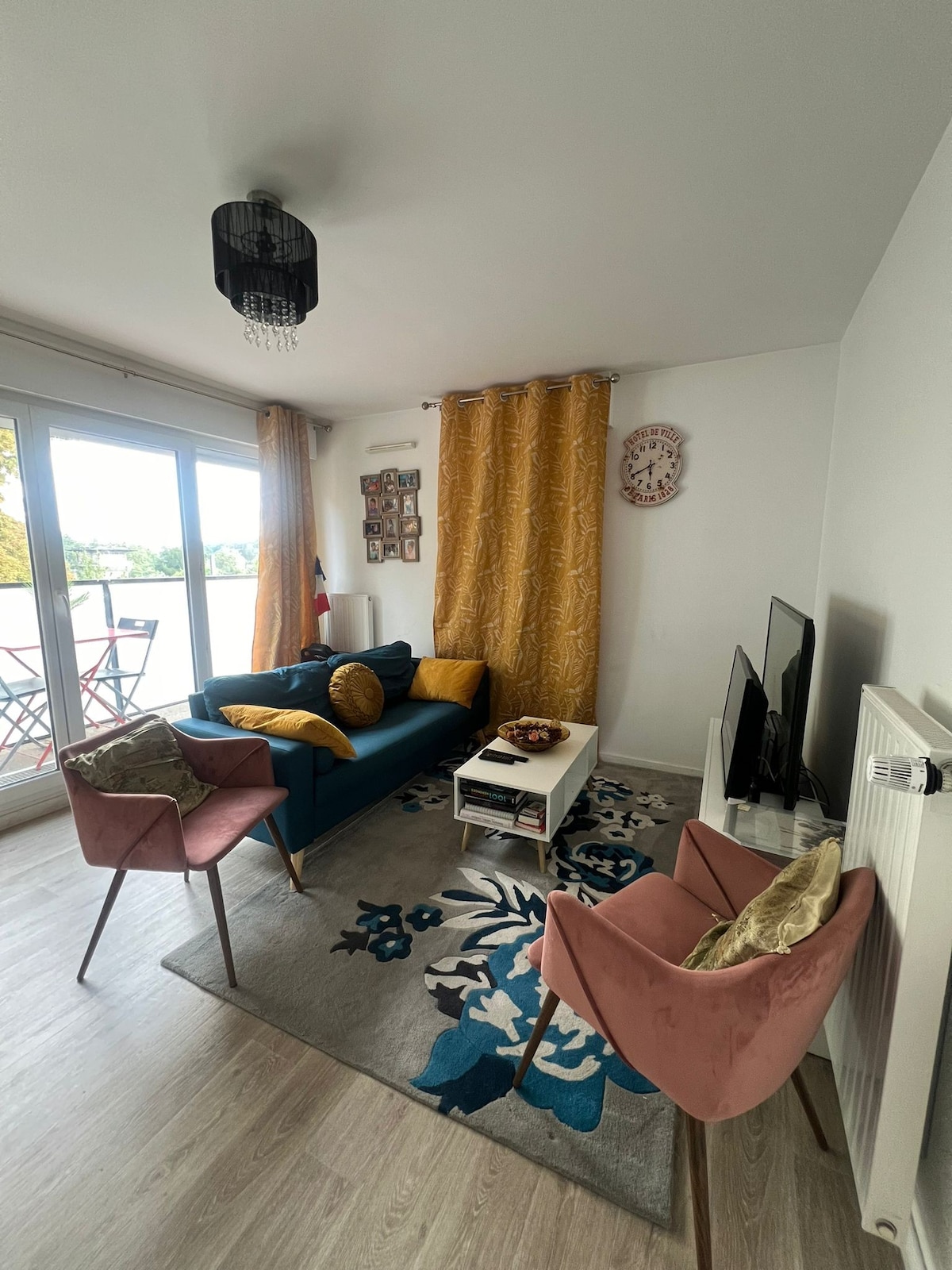 A cozy living area is presented, featuring a blue sofa adorned with decorative cushions. Two pink armchairs are positioned nearby. A coffee table displays items on a patterned rug. Large glass doors open to a balcony, framed by golden curtains.