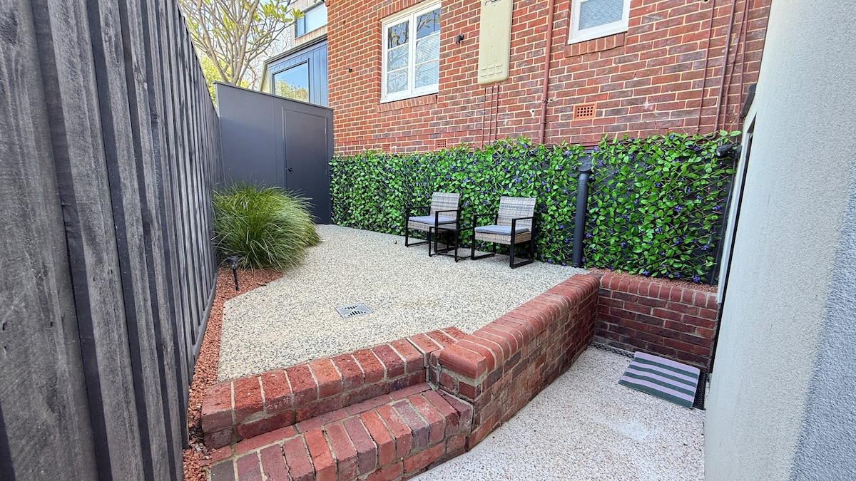 A private outdoor space is enclosed by greenery and features two chairs positioned on a gravel surface. Brick steps lead to the seating area, providing a cozy environment for relaxation.