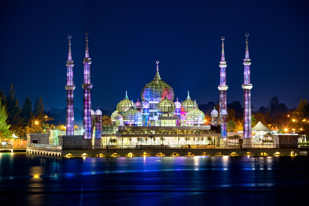 A stunning architectural structure is illuminated at night, featuring multiple domes and tall spires adorned with vibrant colors. The building reflects beautifully on the water, surrounded by softly glowing lights that enhance its grandeur against the night sky.