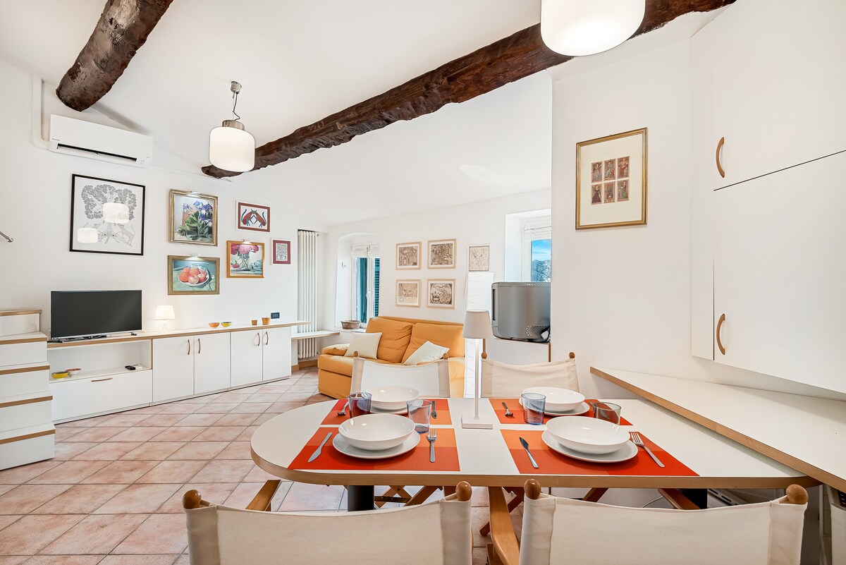 A cozy living area features a vibrant orange sofa and a white dining table set for four. Artistic wall decorations add character, while a television is positioned nearby. Exposed wooden beams grace the ceiling, and natural light fills the space through large windows.