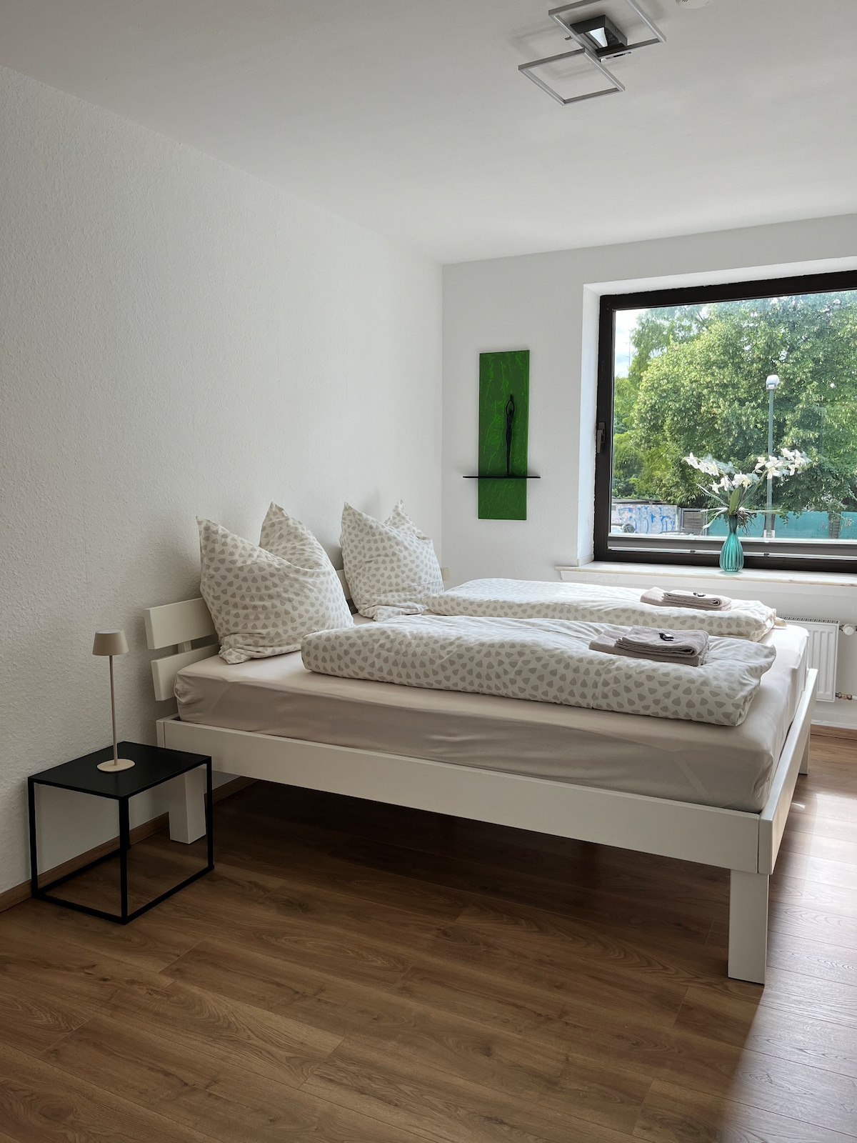 A light-filled sleeping area features two spacious beds with patterned bedding. A small nightstand is positioned beside one bed. A large window overlooks green trees outside, allowing natural light to create a bright and airy atmosphere.