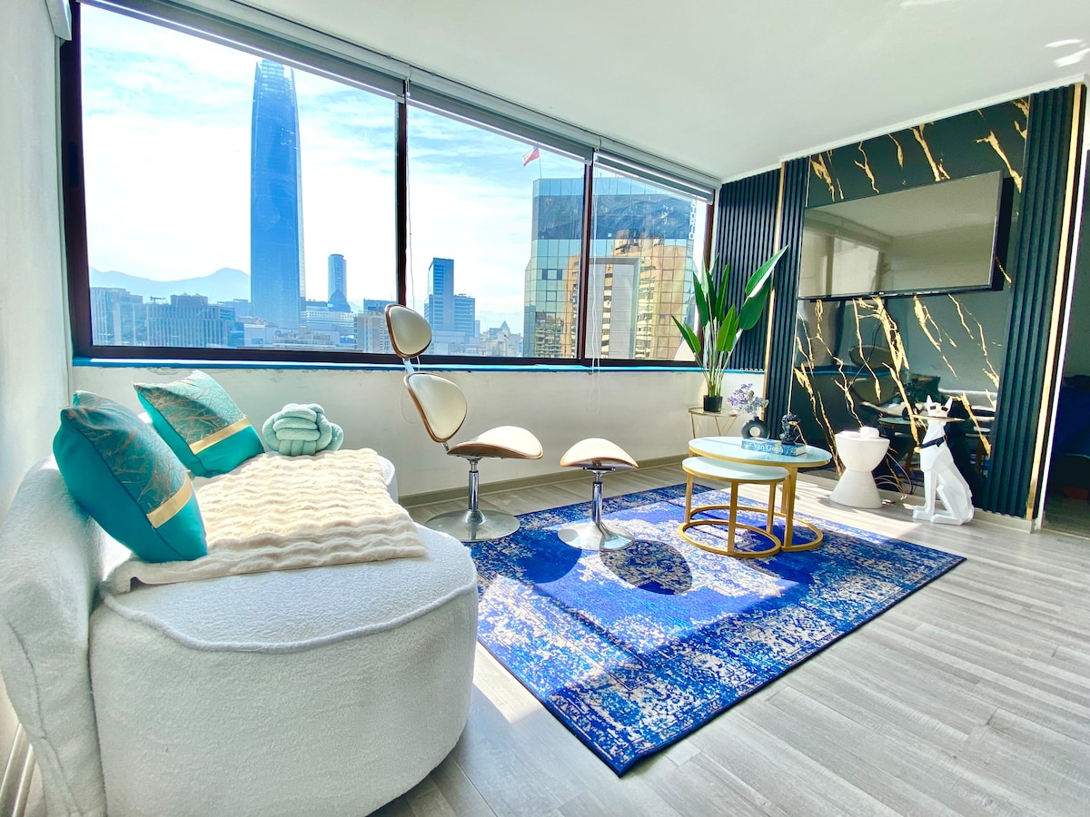 A bright living area is positioned next to large windows, offering a panoramic view of the iconic Torre Costanera. A plush white seating area complements a colorful rug and a small round table. Modern chairs add a stylish touch to the inviting space.