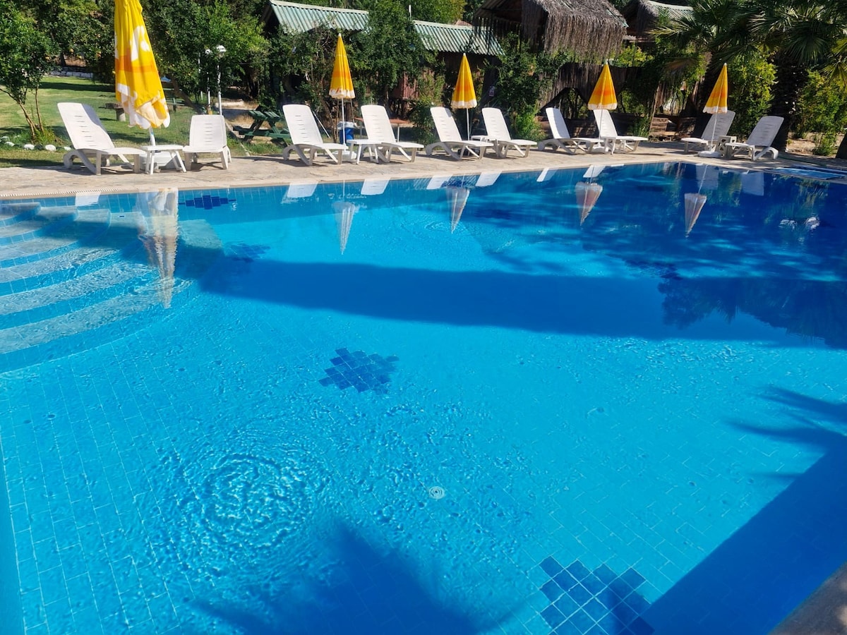 A clear swimming pool reflects sunlight, surrounded by lounge chairs with yellow umbrellas. The pool's mosaic tiles create an inviting pattern, while the lush greenery in the background adds a natural ambiance.