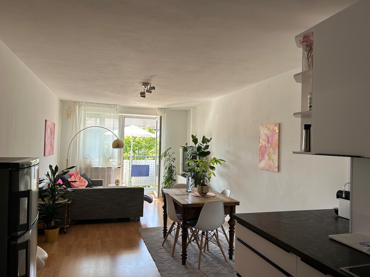 A spacious living area is shown, featuring a comfortable couch and a dining table with chairs. Large glass doors provide access to a sunny balcony. Potted plants are present, adding greenery, while soft natural light illuminates the room, enhancing the calm decor.