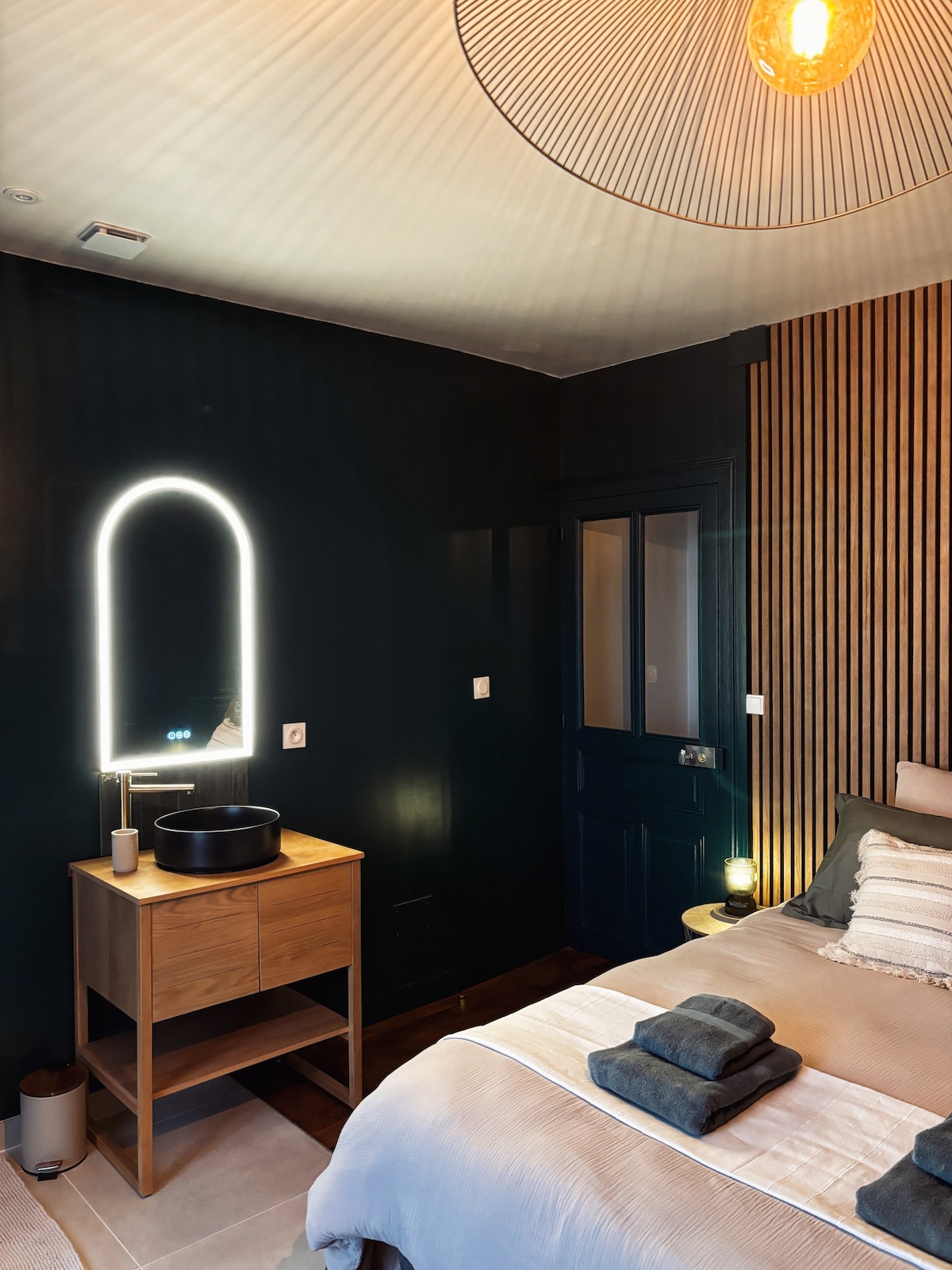 A stylish bedroom features dark green walls and wooden accents. A spacious bed is adorned with neatly arranged linens. A round mirror with integrated lighting and a wooden nightstand accompanied by a round basin are visible, creating a modern and functional ambiance.