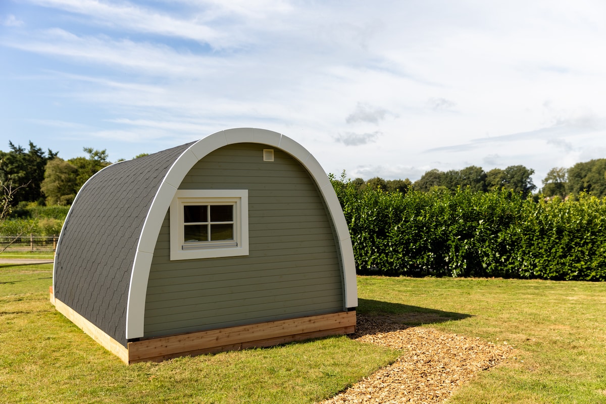 A charming arched structure sits in a grassy area, surrounded by neatly trimmed hedges. A small gravel path leads up to the building, which features a single window and a door on one side, offering a glimpse of its inviting design.