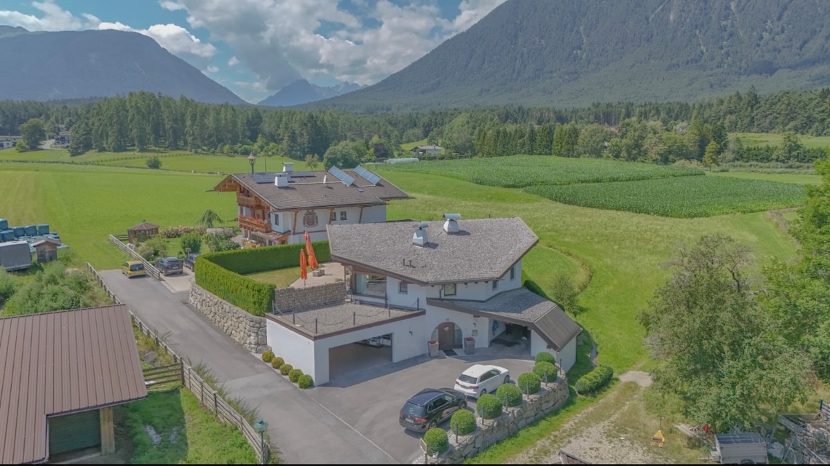 An aerial view of the apartment complex is captured, surrounded by lush green fields and towering mountains. The modern building features a stone façade and a triangular roof, with red accents visible on the adjoining structure. Large windows reflect the natural landscape, highlighting the serene setting.