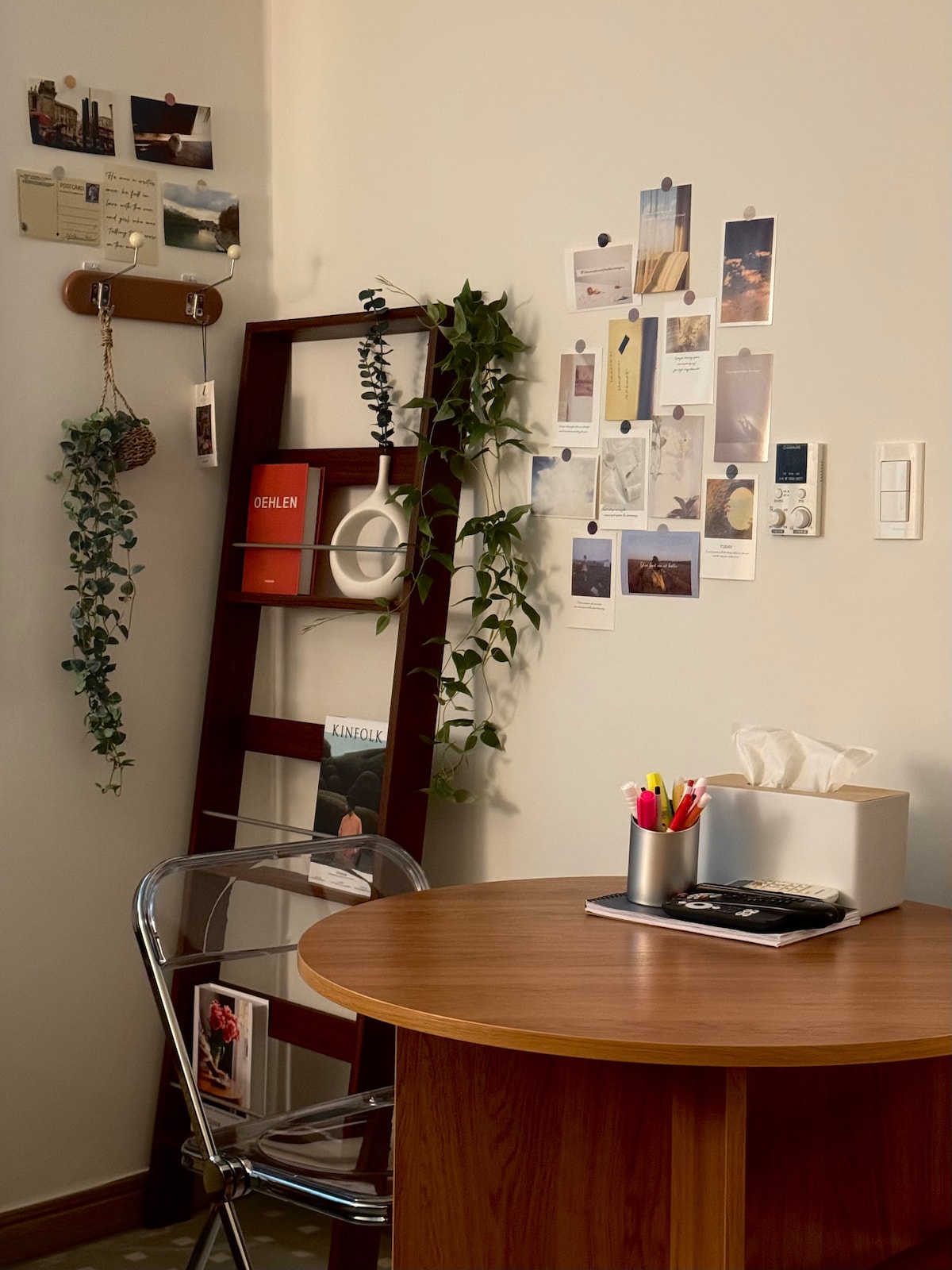 A corner workspace features a circular wooden table beside a clear acrylic chair. A wooden ladder shelf displays various photographs and decorative items, while green plants are draped alongside. A stationery holder with pens and tissues sits on the table, creating an organized atmosphere.