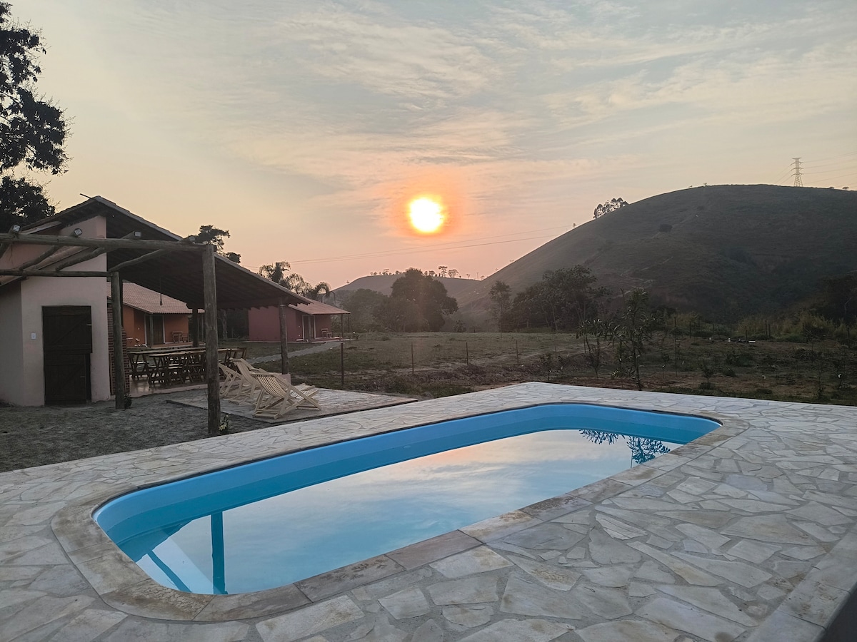 A serene pool reflects the warm colors of a sunset, surrounded by natural landscapes. The distant hills and gentle skyline create a calming backdrop, while rustic buildings are seen nearby, providing a sense of place within the tranquil setting.