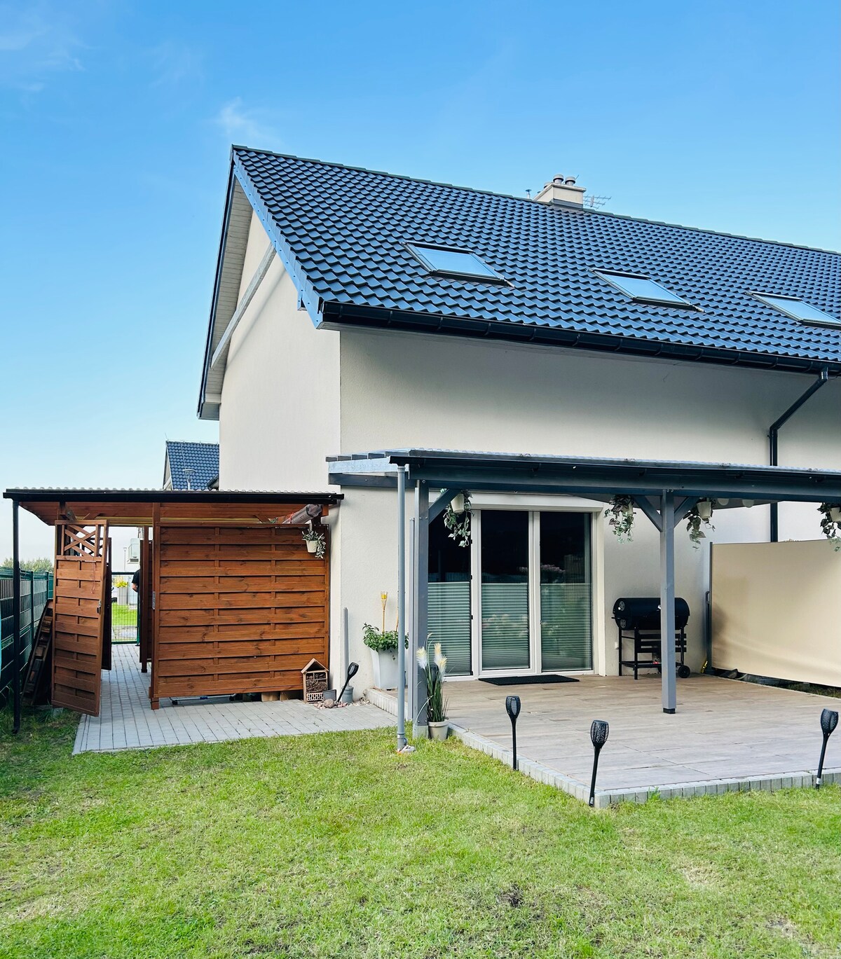 The exterior features a well-maintained garden area with a wooden deck and a covered seating space. A large window offers a view into a bright indoor space, and the building features a contemporary roof design, enhancing the overall architectural appeal.