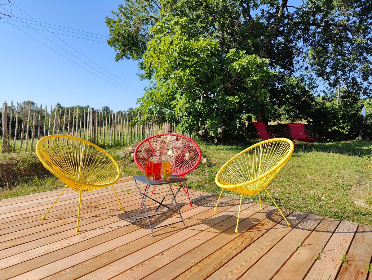 A wooden deck features three colorful chairs: two in yellow and one in red, arranged around a small table. The scene is complemented by a lush green lawn and trees in the background, creating a relaxed outdoor space for gatherings.