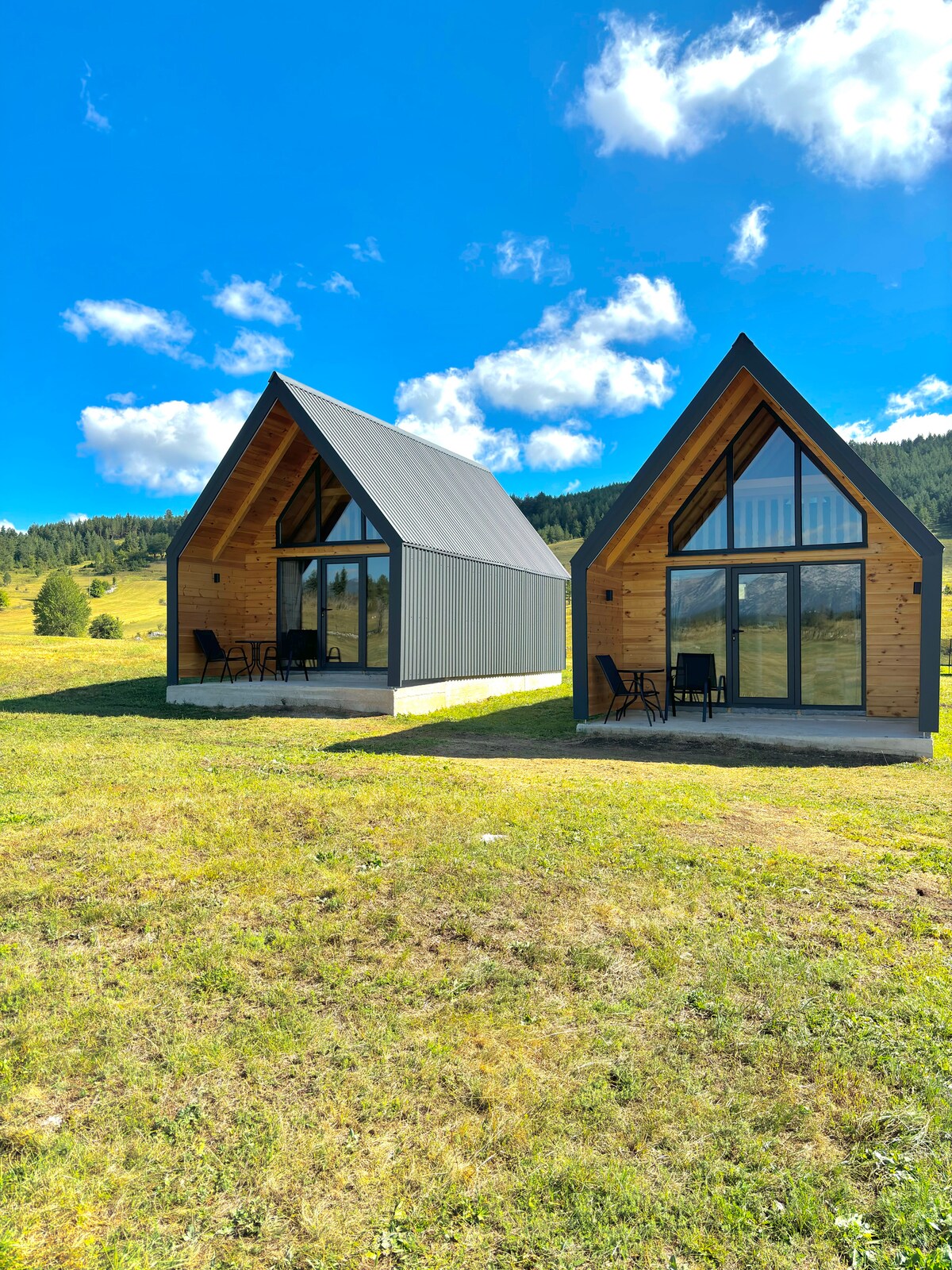 Two glamping cabins are set in a meadow, featuring large glass doors that open to the outdoors. The structures have sloped roofs and wooden accents, with seating areas located on covered patios. The surrounding landscape includes rolling hills and patches of greenery under a clear blue sky.