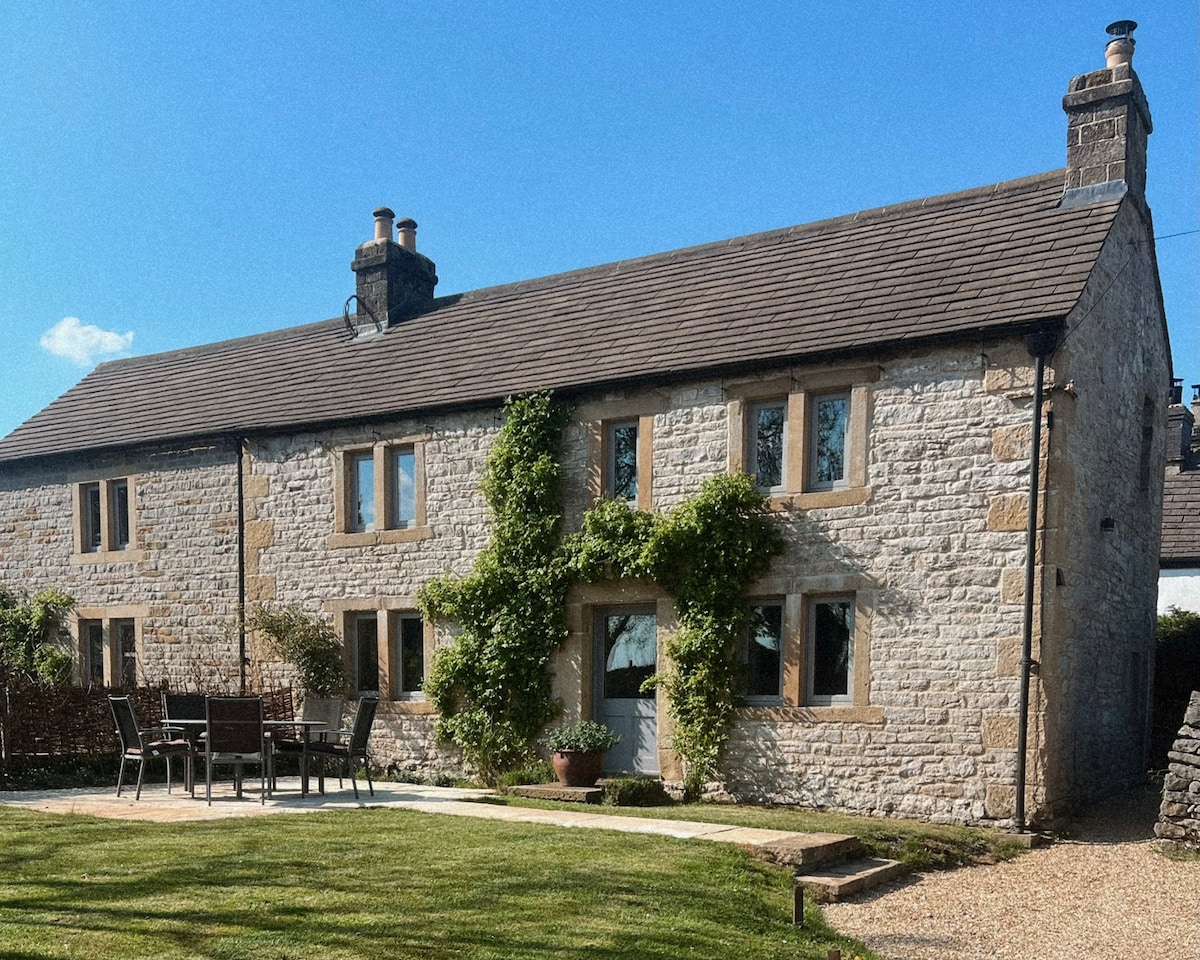 The stone cottage features a charming façade adorned with climbing greenery. A patio area with outdoor furniture is positioned in front of the cottage, surrounded by a well-maintained lawn. Large windows invite natural light, while the property is set against a backdrop of clear blue skies.