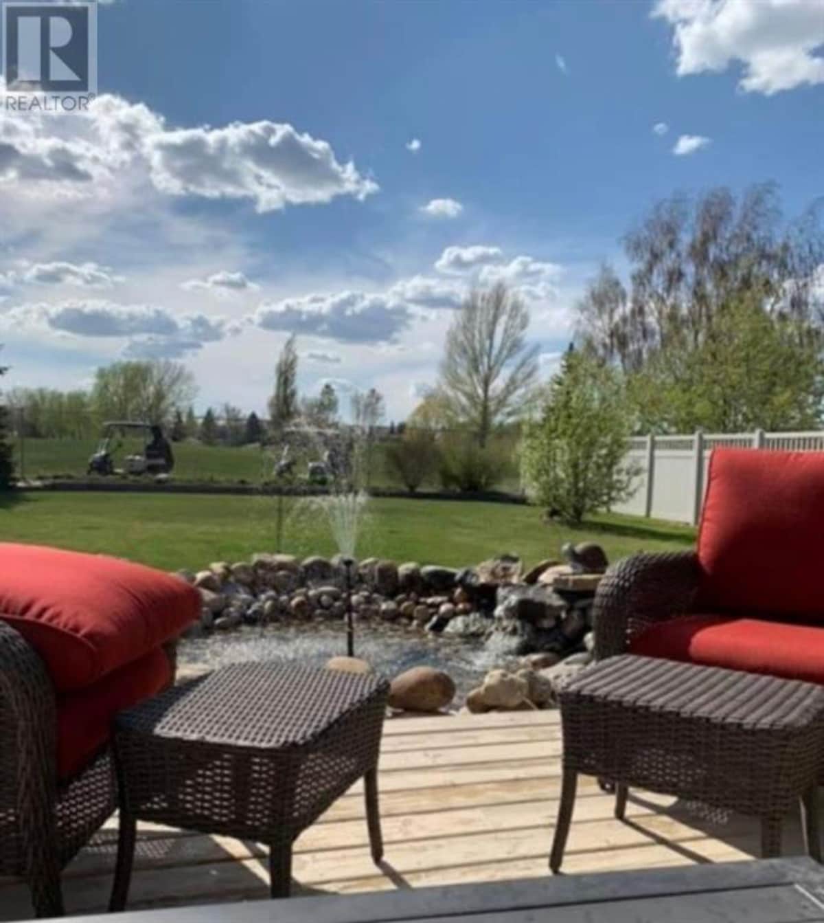 A serene outdoor space is displayed, featuring two woven armchairs with bright red cushions. A small water feature, surrounded by decorative stones, creates a focal point. Lush green grass and trees extend in the background under a blue sky dotted with fluffy white clouds.