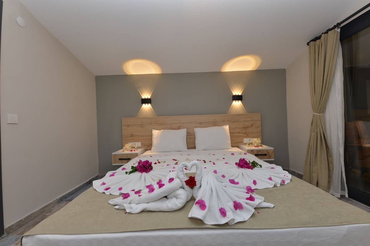 A calming bedroom features a spacious bed adorned with white linens and decorative floral arrangements. Artisan-crafted swan towel displays enhance the inviting atmosphere. Soft lighting from wall sconces creates a serene ambiance, while large windows are dressed with light curtains, allowing for natural light.