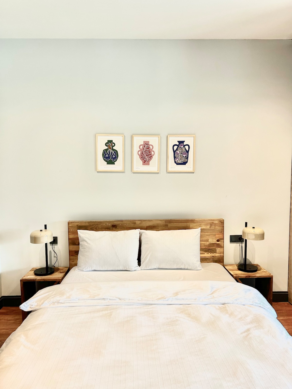 A comfortable sleeping area features a wooden headboard with two light-colored pillows. On either side, wooden nightstands with stylish lamps are positioned. Above the bed, three framed art pieces depicting colorful vases add a decorative touch to the neutral-colored wall.