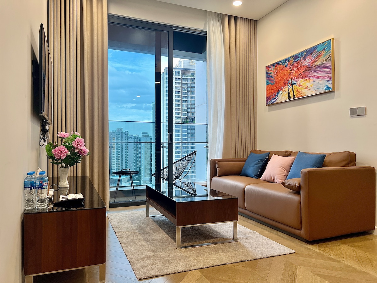 A modern living room features a brown leather sofa adorned with decorative cushions in blue and pink. A glass coffee table sits on a soft area rug, while large windows offer a scenic view of the cityscape. Fresh flowers are displayed on a side table.