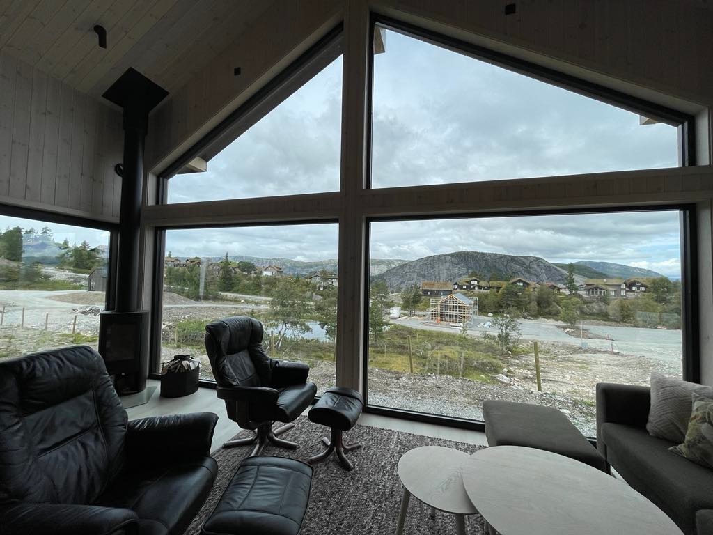 Large windows provide a panoramic view of the surrounding landscape, featuring mountains and valleys. Comfortable seating is arranged in a cozy living area, with a rug and coffee tables enhancing the space. Natural light floods the room, creating an inviting ambiance.