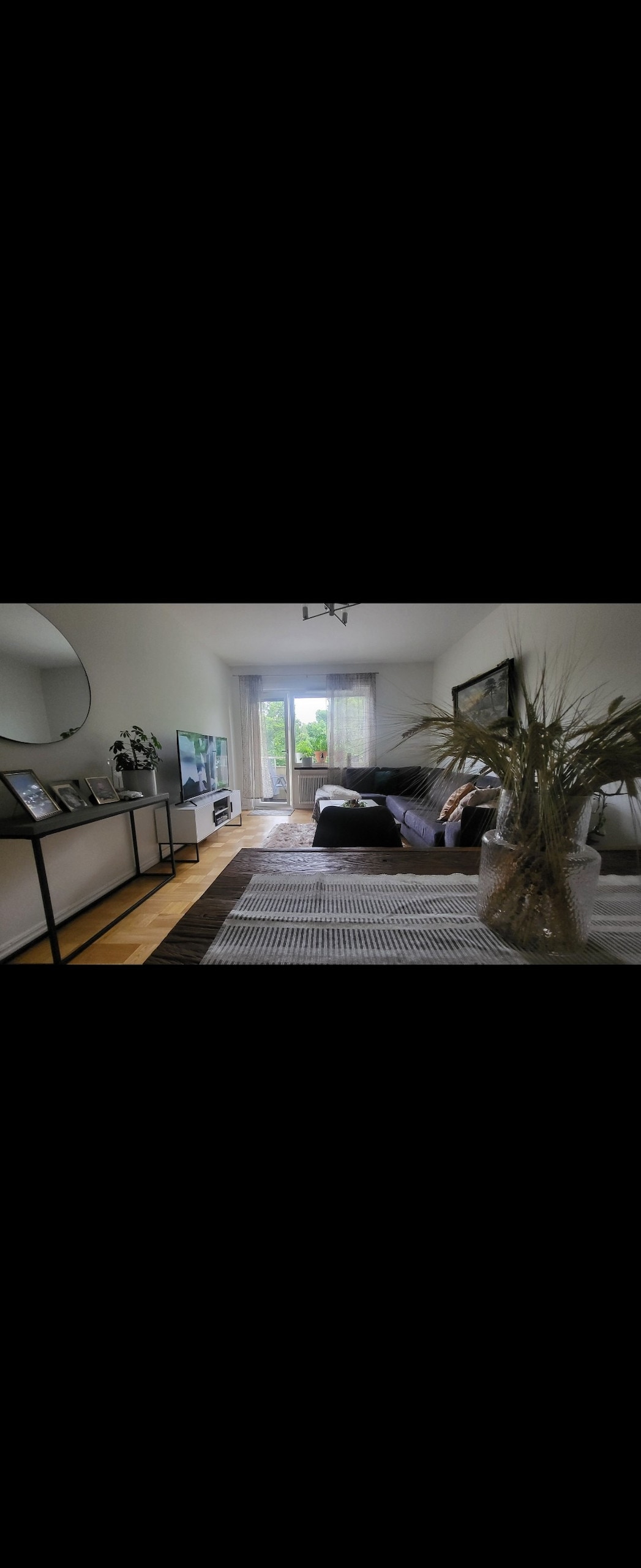 A living area features a spacious sofa facing a wall-mounted television. A round mirror is displayed above a sleek console table. Natural light filters through sheer curtains, highlighting the texture of a patterned rug and a decorative plant in a vase.