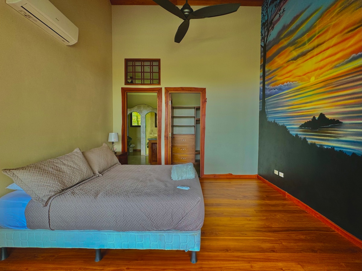 A spacious room features a large bed with a neutral-toned blanket. A colorful mural depicting a sunset scene adorns one wall. A ceiling fan provides ventilation, and a soft light fixture is near the bedside. A doorway leads to a private bathroom.